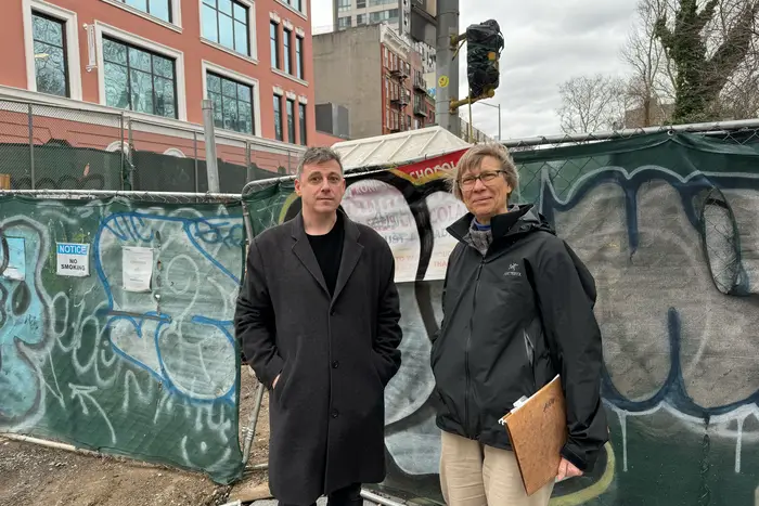 A man and woman stand in front of construction site fencing covered in graffiti.